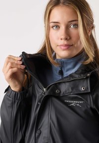 Czarna wodoodporna kurtka z zamkiem błyskawicznym i zapięciem na zatrzask, z logo, elastycznymi mankietami i podszytym kołnierzem. Noszona na jasnoniebieskim polarku.