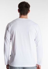 Man with short brown hair wearing a plain white long-sleeve shirt and gray pants, viewed from the back against a white background.