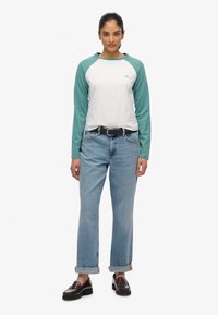 Langermige raglan t-shirt in wit en teal met een ronde hals; gecombineerd met lichtblauwe rechtvallende jeans en bruine instappers.