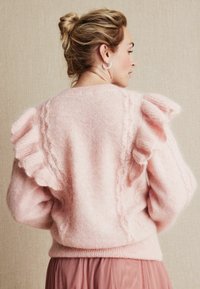 Jasnoróżowy sweter z dzianiny z falbanami na ramionach, teksturowanym wzorem na plecach oraz ściągaczem u dołu.