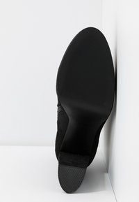 Semelle d'une chaussure à talon noire appuyée contre un mur blanc, montrant la surface inférieure lisse avec un embout de talon texturé.