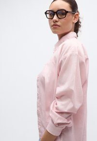 Lys rosa skjorte med lange ermer og knapper, med oppbrettede mansjetter. Modellen har på seg store briller med mørk ramme. Glatt stofftekstur, løs passform.