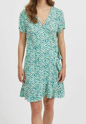 Vrouw in een kortmouw, wikkeljurk met een groen, wit en blauw bloemenpatroon, V-halslijn en een gerimpelde zoom.