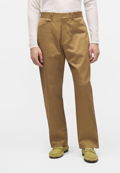 Homme portant un pantalon droit marron clair, des mocassins vert clair et une chemise blanche à manches longues, debout devant un fond uni.