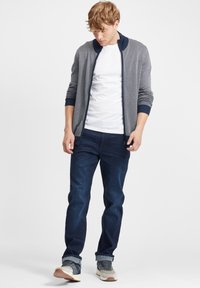 Cárdigan ligero en gris con un patrón texturizado, combinado con pantalones vaqueros azul oscuro. Con cierre de cremallera y cuello redondo. Zapatillas casuales.