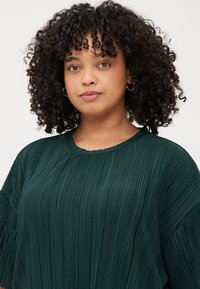 Μια νεαρή γυναίκα με σγουρά μαλλιά φοράει μια πράσινη πλισέ μπλούζα, κοιτάζοντας απευθείας στην κάμερα με ουδέτερη έκφραση απέναντι σε απλό φόντο.