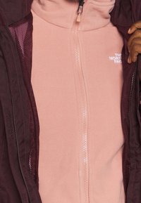 Bessenkleurige buitenjas over een lichtroze fleece met rits. Voorzien van een hoge kraag, ritssluiting en merkopdruk op de fleece.