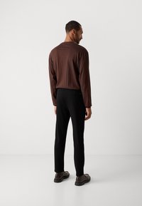 Hombre vestido con una camisa marrón de manga larga, pantalones negros y zapatos marrones oscuros, de pie frente a una pared blanca lisa.