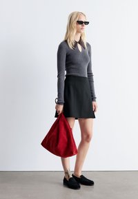 Grauer gerippter Strickoberteil mit Schlüssel-Ausschnitt, schwarzer ausgestellter Rock, schwarze Loafer und eine große Handtasche aus rotem Wildleder. Glatte Textur, zeitgenössisches Design.