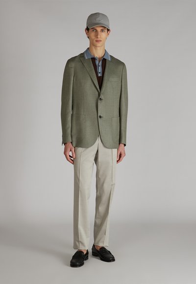 Młody mężczyzna w szarym czapce, zielonym żakiecie, niebiesko-brązowej koszulce polo, beżowych spodniach i czarnych mokasynach stoi na tle jednolitym.