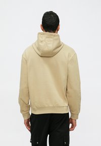 Felpa beige con una vestibilità aderente, dotata di un grande cappuccio, polsini e orlo a coste. Il tessuto sembra morbido con una texture liscia e opaca.