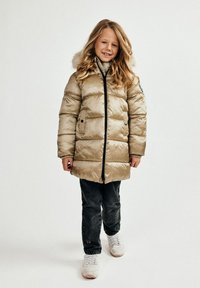 Lys beige puffjakke med pelsforet hætte, vandret quiltning, lynlåse og knaplommer. Båret over en grå sweater og jeans.