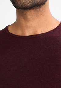 Burgunderfarbene Strick-Crewneck-T-Shirt, das eine weiche Textur und eine figurbetonte Passform zeigt. Der Ausschnitt ist weit und lässt die glatte Haut darunter sichtbar werden.