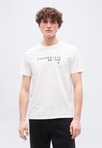 Νέος άνδρας με σγουρά μαλλιά, φοράει ένα λευκό μπλουζάκι Calvin Klein με μαύρο κείμενο και μαύρο παντελόνι, στέκεται μπροστά σε απλό φόντο.