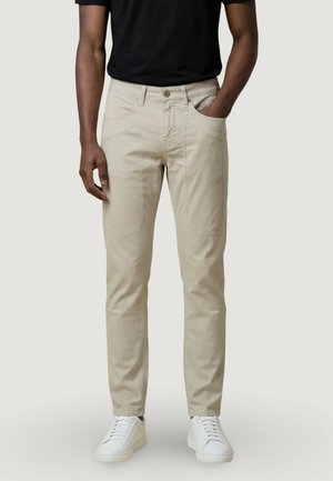 Uomo con pantaloni beige aderenti, maglietta nera e sneaker bianche, in piedi con una mano in tasca su sfondo chiaro.