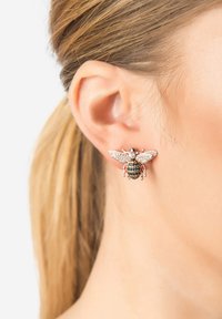 Pendiente con forma de abeja en tonos de plata y oro, con acentos de piedras de strass y alas detalladas, anidado en el lóbulo de una oreja.