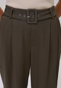Pantalon taille haute en tissu vert olive foncé. Il présente une large ceinture avec une boucle carrée, des œillets et des plis pour plus de texture.