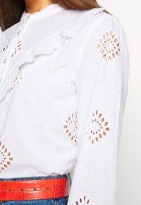 Blouse blanche avec bordure en dentelle et motifs floraux ajourés. Manches longues avec motifs découpés. Associée à une ceinture texturée orange vif.