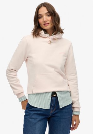 Sweat à capuche rose clair en matériau doux avec un cordon de serrage, poche avant et poignets côtelés. Porté sur une chemise rayée bleue. Jeans bleus.