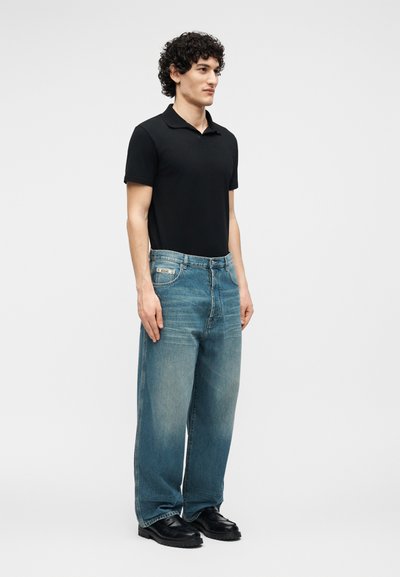 Νεαρός άνδρας με σγουρά μαλλιά φοράει μαύρο πόλο με κοντά μανίκια, χαλαρά ξεθωριασμένα μπλε τζιν και μαύρα παπούτσια, ενώ στέκεται μπροστά σε απλό φόντο.