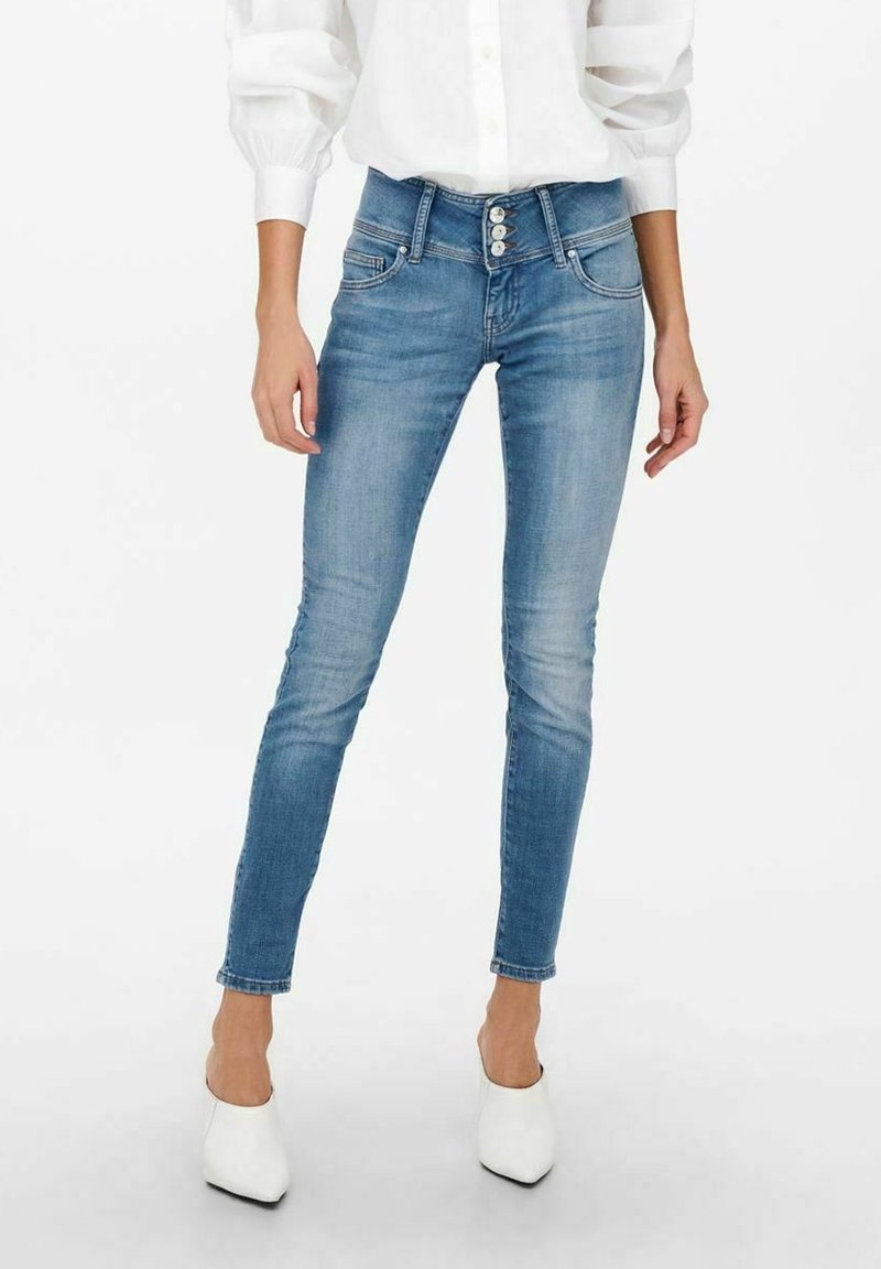 Jean skinny bleu clair usé avec fermeture à triple bouton, associé à des talons blancs à bout pointu et une chemise blanche à manches longues.