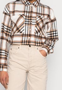Koszula z flaneli w kratę w kolorach białym, brązowym i czarnym, z dwoma kieszeniami na piersi i zapięciem na guziki, zestawiona z jasnobeżowymi, wysokim stanem spodniami.