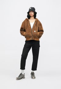 Uma mulher vestindo um hoodie marrom com fecho, chapéu tipo bucket preto, calças pretas de punho, meias brancas e sapatos de caminhada cinzentos, de pé com as mãos nos bolsos.