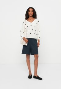 Maglione bianco a maglia con motivi a cuore neri, pantaloni shorts blu navy e scarpe slip-on nere. Gli accessori includono una piccola borsa chiara.