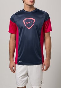 Nike Dri-FIT urheilusuoja, jossa on tummansininen vartalo ja vaaleanpunaiset lyhyet hihat sekä logo, valmistettu kevyestä ja kosteutta siirtävästä kankaasta.