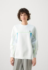 Sweatshirt blanc avec des accents bleus et verts, poignets côtelés, col rond et logo brodé. Le matériau semble doux et texturé.