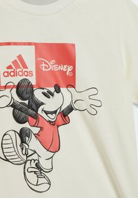 Maglietta color crema con un grande Mickey Mouse ricamato di nero, vestito con una maglietta rossa, e i loghi di Adidas e Disney su uno sfondo rosso.