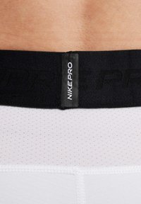 Fascia elastica nera con etichetta "NIKE PRO" su tessuto bianco testurizzato con piccoli punti. Il capo è aderente e si allunga leggermente.