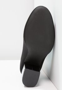 Semelle et talon d'une botte noire à la cheville reposant contre un coin blanc, montrant la bande de roulement et la surface lisse.