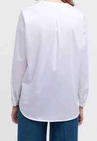 Blouse blanche à manches longues avec un col arrondi, un pli au dos et une texture lisse. Comprend des poignets ajustés et un ourlet incurvé.