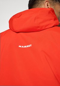 Veste rouge avec une texture lisse, dotée d'une capuche et d'un logo blanc « MAMMUT » bien visible à l'arrière. Design léger pour les activités de plein air.