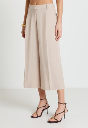 Pantaloni beige a gamba larga e taglio corto abbinati a sandali con tacco alto scuro e cinturini, indossati da una persona in piedi.