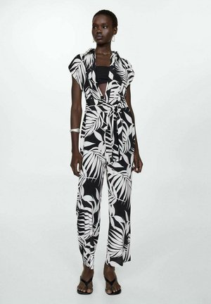 Femme portant une combinaison à imprimé feuillage noir et blanc avec des manches courtes, une taille cintrée, un top tube noir, un bracelet en argent et des talons ouverts noirs.