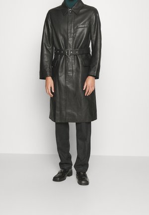 Uomo con trench coat di pelle nero con cintura, pantaloni scuri e scarpe nere in piedi davanti a sfondo bianco.