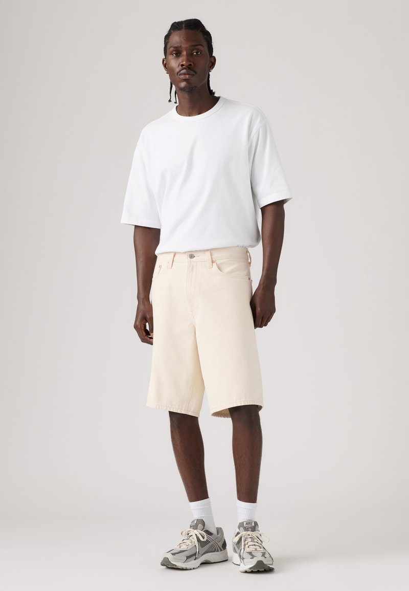 Uomo con capelli intrecciati, che indossa una t-shirt bianca, pantaloncini beige al ginocchio, calzini bianchi e scarpe da ginnastica grigie, in piedi su uno sfondo neutro.