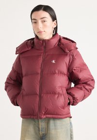 Giacca a vento bordeaux con colletto alto, chiusura con zip e bottoni a pressione sul cappuccio. Presenta un logo sul petto e polsini a costine.