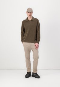 Olivgrüner, gestrickter Pullover mit Kragen, beigen Chinos und schwarzen Sneakers, ergänzt durch eine hellbeige Mütze.