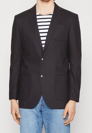 Mężczyzna w czarnej dwurzędowej marynarce, granatowo-białej koszuli w paski oraz jasnoniebieskich dżinsach, pokazany od szyi do talii, stojący na białym tle.