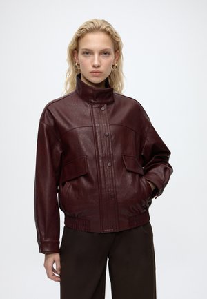 Frau mit blondem Haar trägt eine dunkelbraune Lederjacke mit hohem Kragen und Knöpfen sowie eine dunkle Hose und steht vor einem einfarbigen hellen Hintergrund.