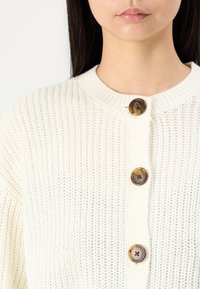 Cardigan blanc en tricot avec un col rond, doté de trois boutons marron à l'avant. Tissu texturé avec une coupe ample.