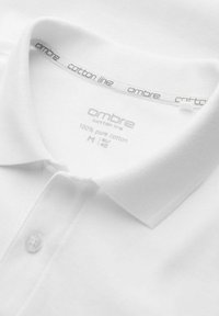 Vit bomullspolo med veckad krage, som har en enkel knapp och en etikett som anger "100% ren bomull" och storlek "M."