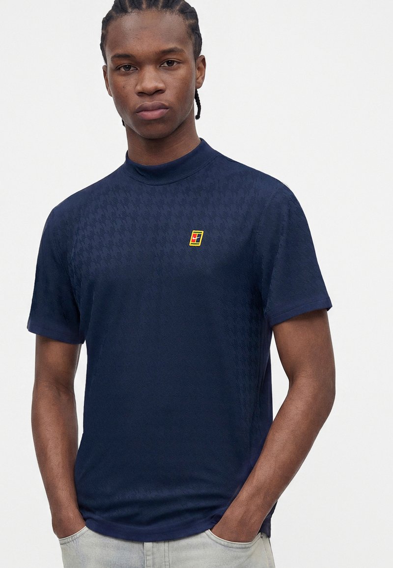 Young man with braided hair wearing a navy blue short-sleeve shirt with a small yellow and red logo on the chest, hands in pockets.
