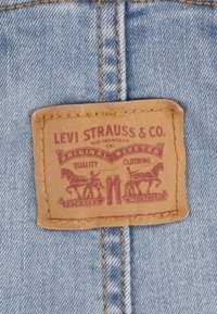 Brun läderlapp med präglad röd text och grafik, som visar två hästar och ett par byxor. Fäst vid ljusblå denim.