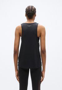 Μαύρο αθλητικό τοπ χωρίς μανίκια με στρογγυλή λαιμόκοψη και λεία υφή. Έχει τύπωμα λογότυπου στην πίσω πλευρά. Φοριέται με ταιριαστά μαύρα κολάν.