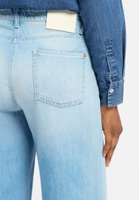 Hellblaue Jeans aus Denim verfügen über eine hintere Tasche mit einem goldenen Nieten und einem weißen Etikett am Bund, begleitet von einem langärmeligen Denim-Hemd.