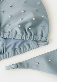 Haut et bas de bikini bleu clair avec des embellissements en perles argentées sur un tissu texturé, posés à plat sur un fond blanc.